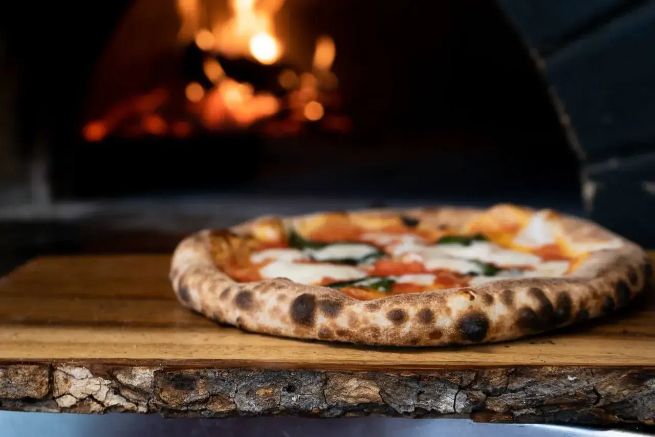 Frisch gebackene Pizza aus Italien, mit geschmolzenem Mozzarella und Basilikum, ruht auf einem Holzbrett vor einem glühenden Ofen.