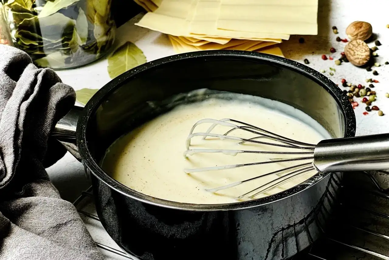Cremige B&eacute;chamelsauce wird in einem Topf mit einem Schneebesen zubereitet. Daneben liegen Lasagneplatten und Gew&uuml;rze.