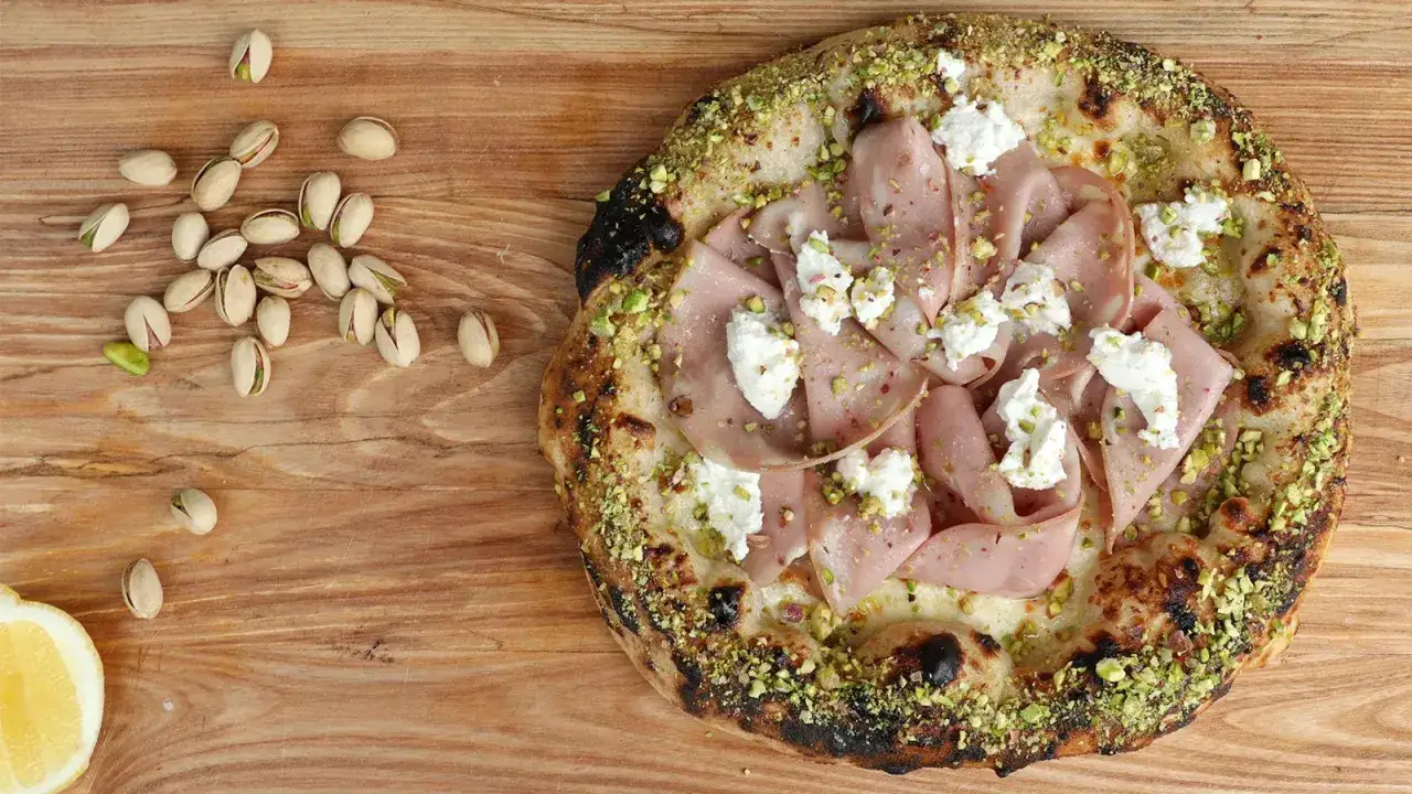 Pizza Mortadella e Pistacchio: knuspriger Teig, Mortadella, Ricotta und gehackte Pistazien. Daneben liegen einige Pistazien und eine Zitronenspalte.