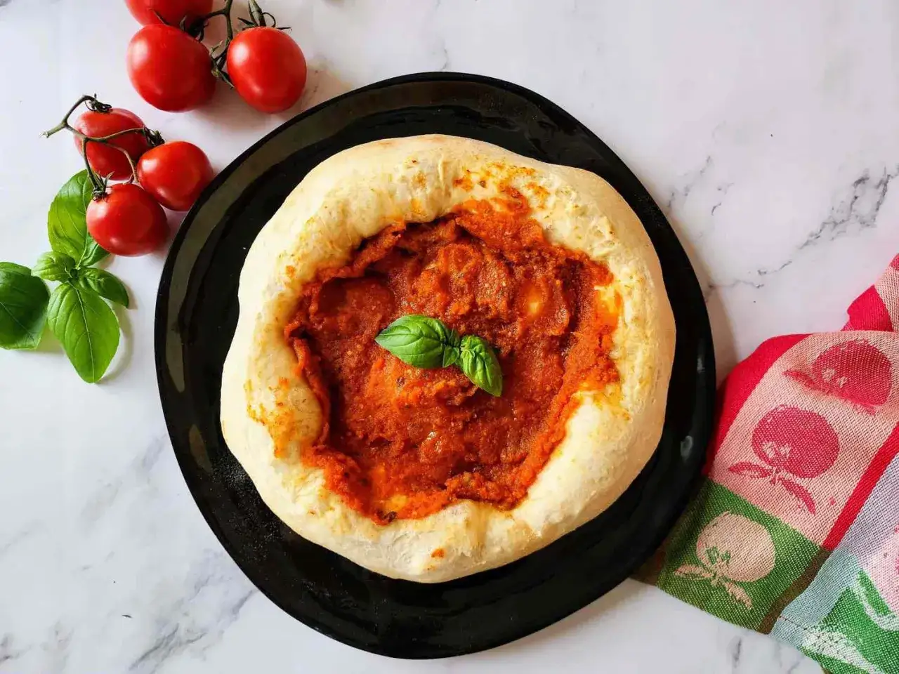 Frisch gebackener Teig mit einer k&ouml;stlichen, original italienischen pizzasauce, garniert mit Basilikum. Daneben frische Tomaten und ein buntes K&uuml;chentuch.