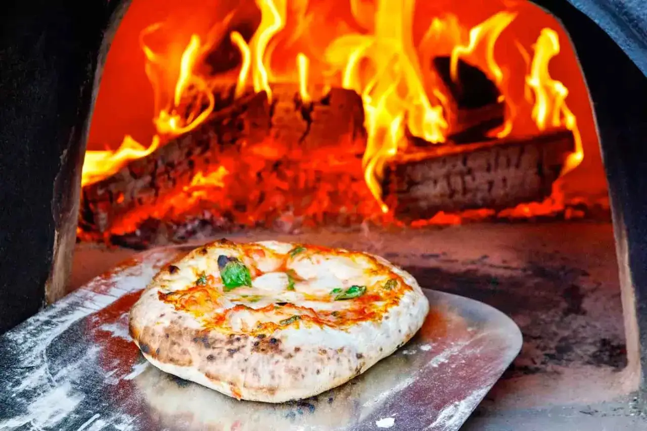 Eine frisch gebackene Pizza Napoli Original auf einem Pizzaschieber vor lodernden Flammen im Holzofen.