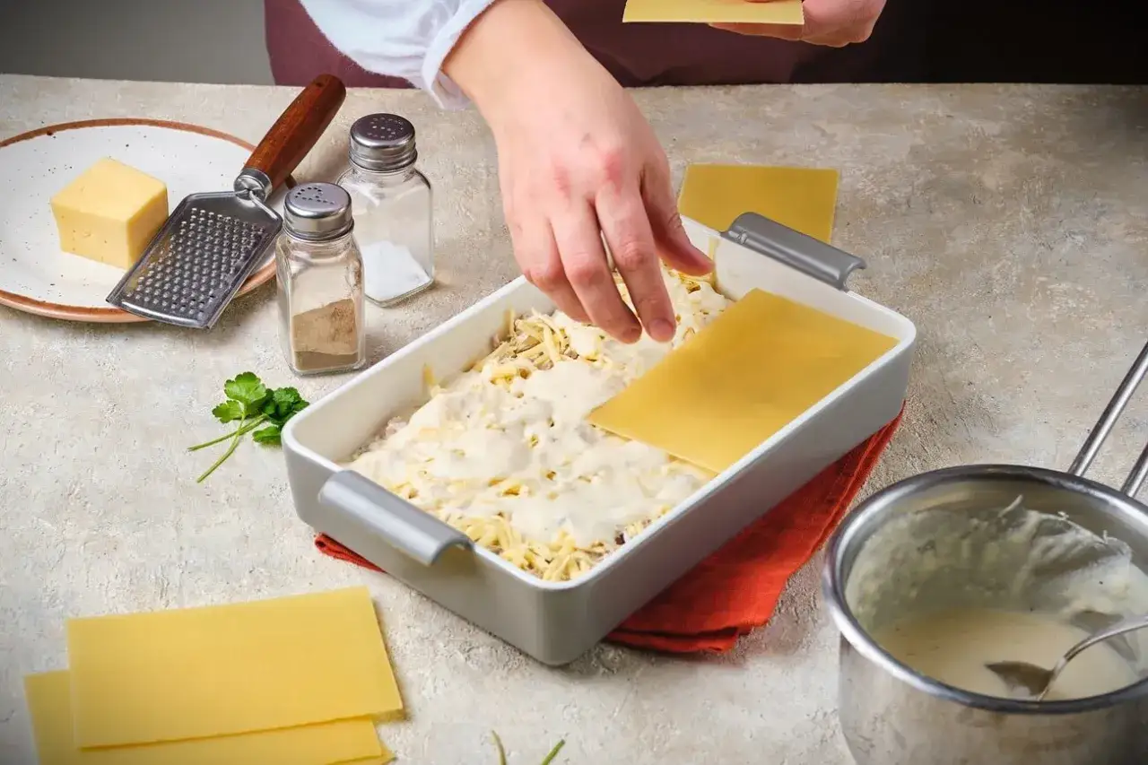 Hände legen Lasagneplatten für ein authentisches italienisches Lasagne Rezept in eine Auflaufform. Käse, Gewürze und Petersilie liegen bereit.