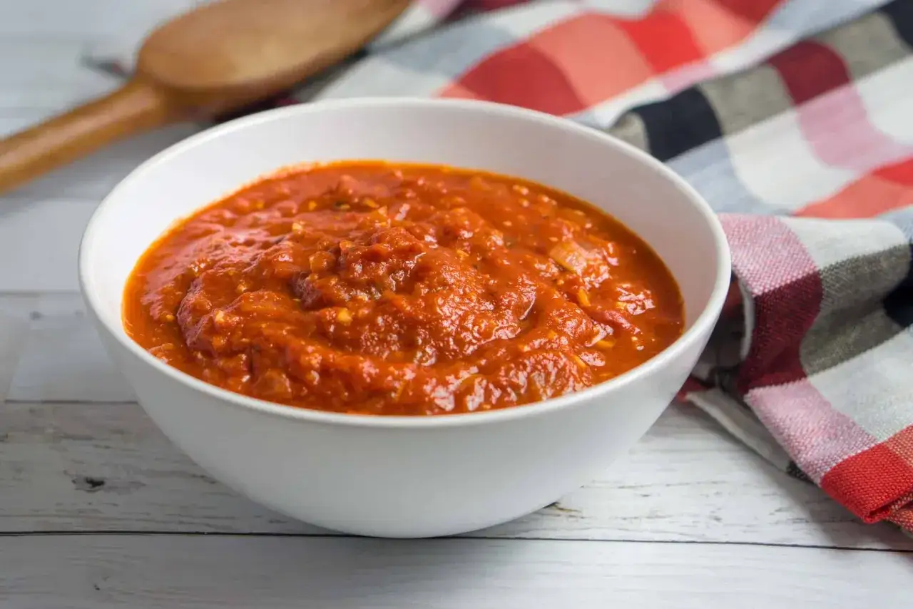 Sch&uuml;ssel mit leckerer San Marzano Pizza Sauce, bereit f&uuml;r die n&auml;chste Pizza. Daneben ein Holzl&ouml;ffel und ein kariertes Tuch.