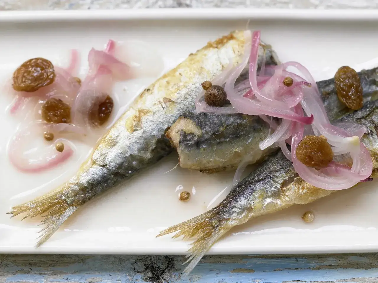 Sardinen in Saor: Gebratene Sardinen mit süß-sauren Zwiebeln, Rosinen und Gewürzen auf einem weißen Teller.