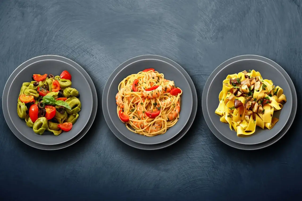 Drei Teller mit italienischen Nudelgerichten: grüne Tortellini mit Tomaten und Oliven, Spaghetti mit Garnelen und Tomaten, Bandnudeln mit Pilzen.