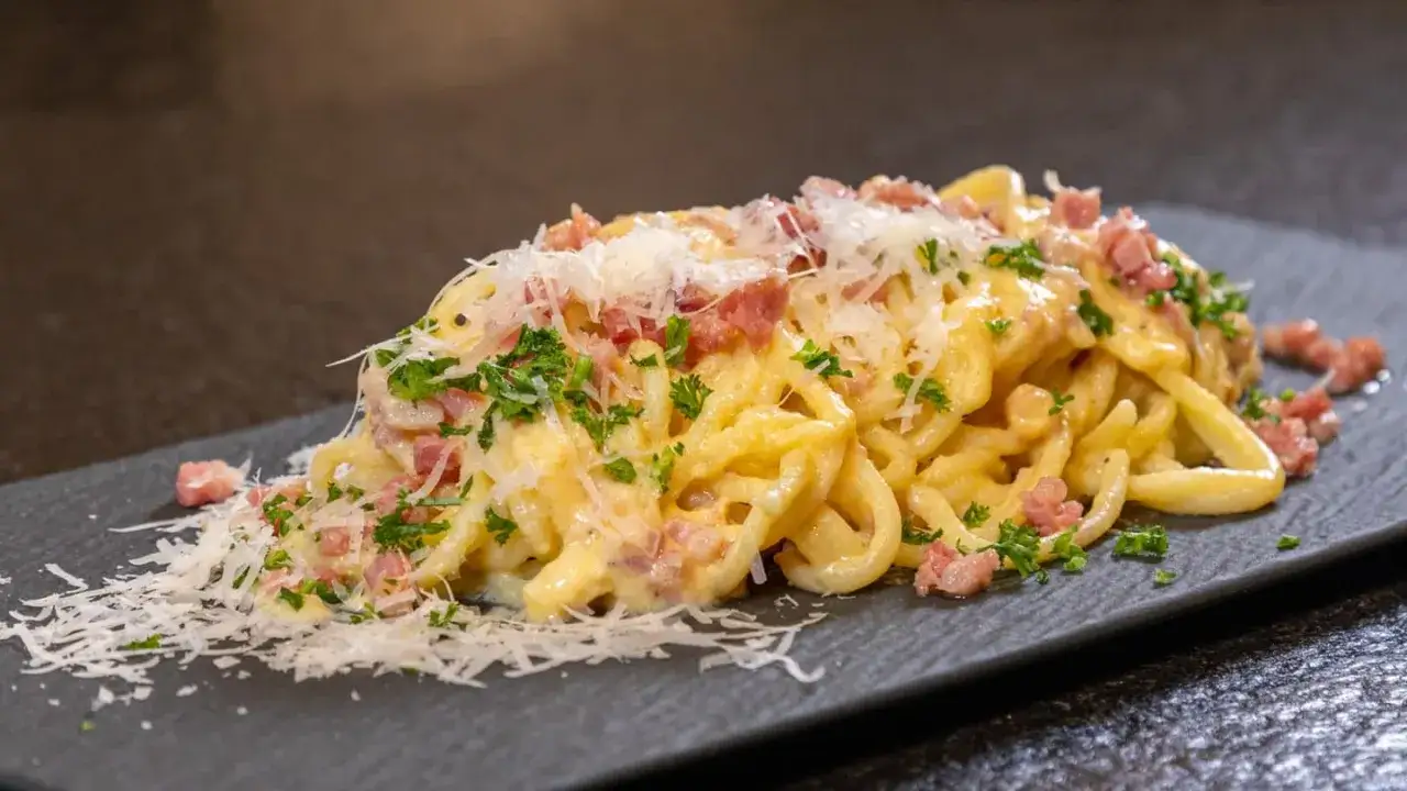 Ein Teller cremiger Pasta Carbonara, garniert mit Speckw&uuml;rfeln, Parmesan und Petersilie, serviert auf einem dunklen Stein.