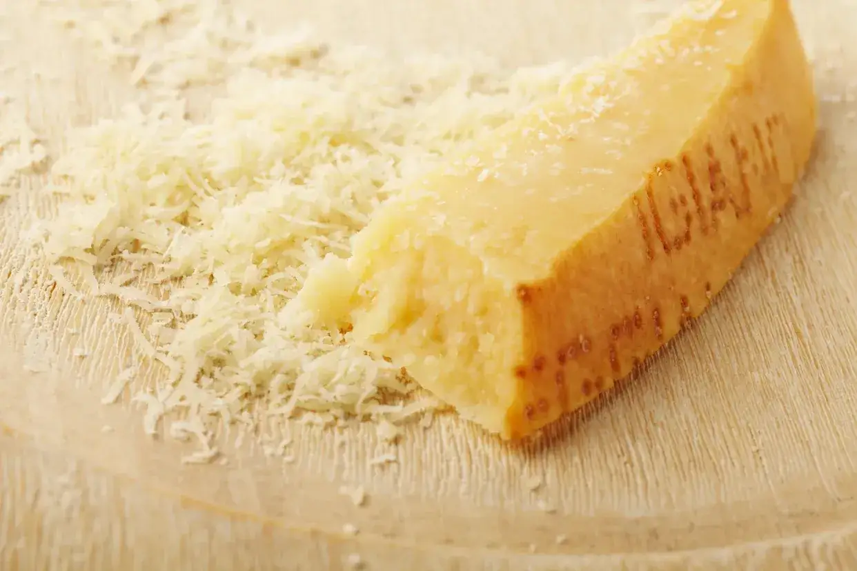 Ein St&uuml;ck Parmesan mit geriebenem K&auml;se daneben. Die Rinde des Parmesans ist mit Pr&auml;gungen versehen.