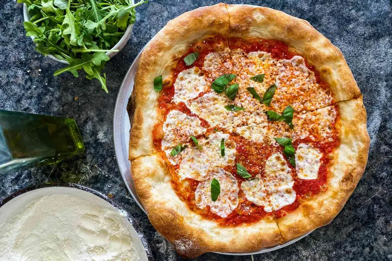 Eine frisch gebackene Pizza Margherita mit Tomatensoße, Mozzarella und Basilikum. Daneben eine Schüssel Rucola, eine Flasche Olivenöl und eine Schüssel Mehl.