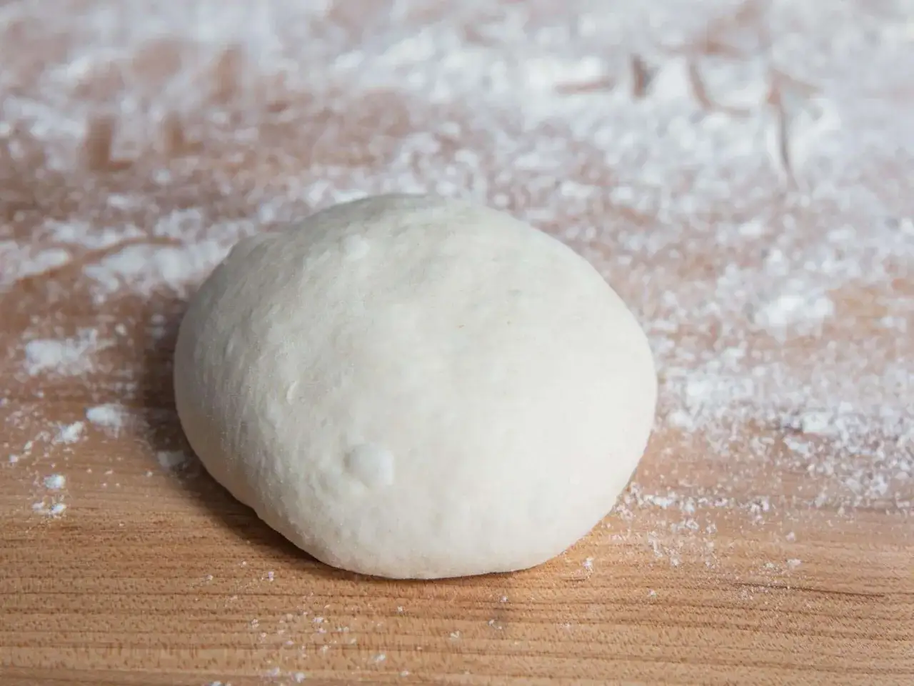 Ein Klumpen frischen Teigs für ein Pizzateig Rezept liegt auf einem Holzbrett, bestäubt mit Mehl.