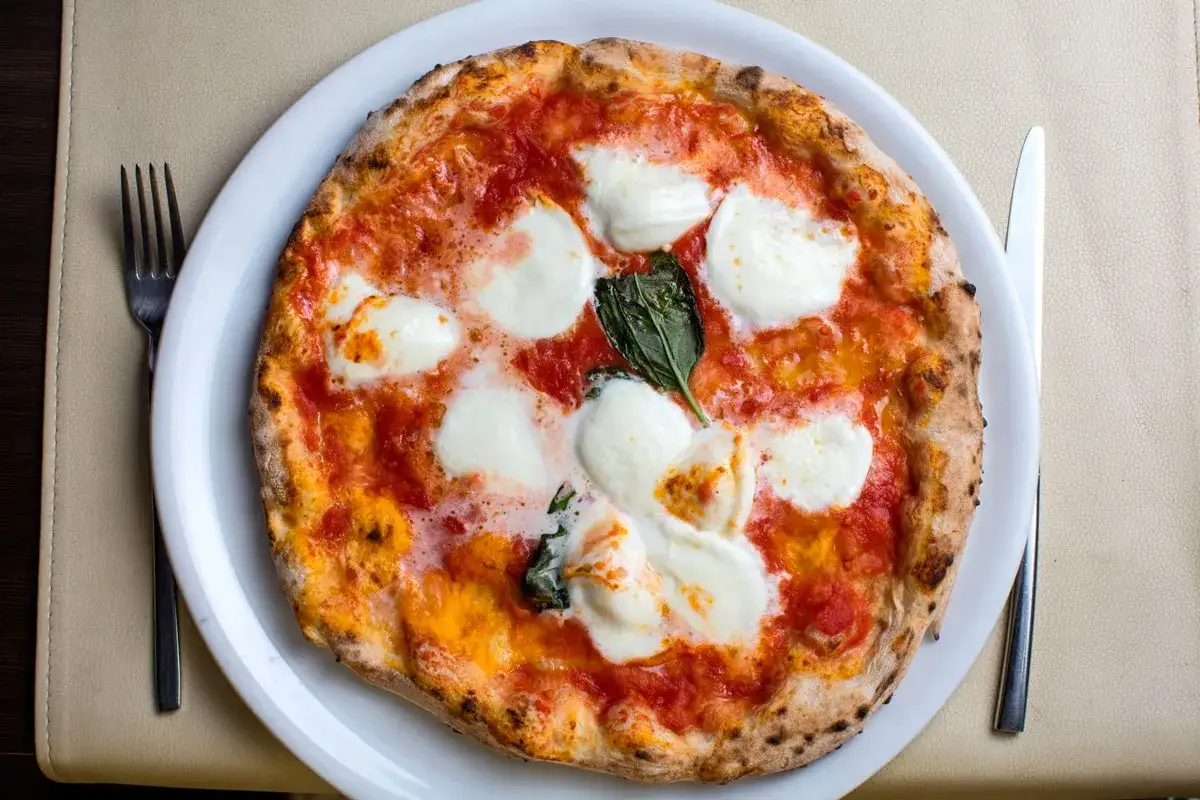 Frisch gebackener Pizzateig f&uuml;r 4 Personen, belegt mit Tomatensauce, Mozzarella und Basilikum. Gabel und Messer liegen bereit.