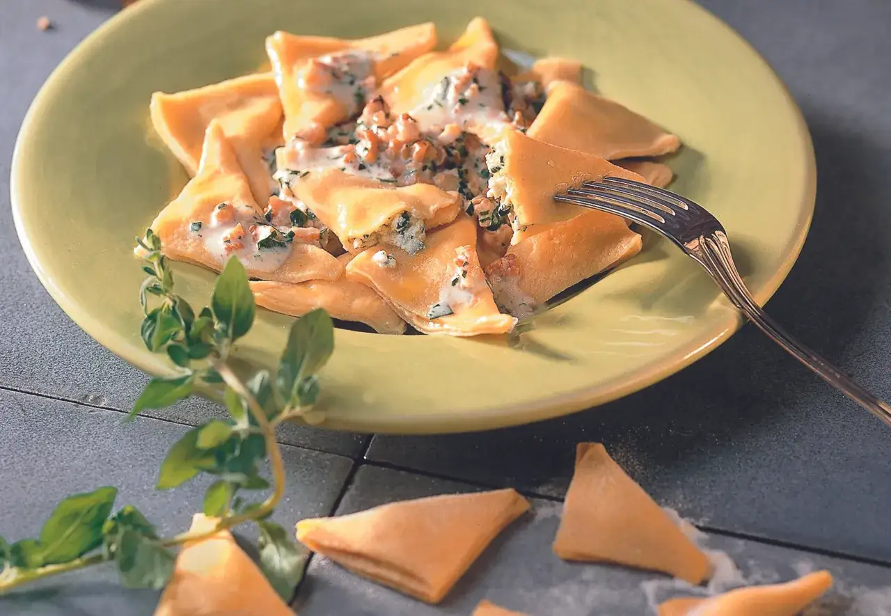 Dreieckige Ravioli mit cremiger Walnuss-Sauce italienisch, garniert mit Kräutern. Eine Gabel nimmt ein Stück auf.
