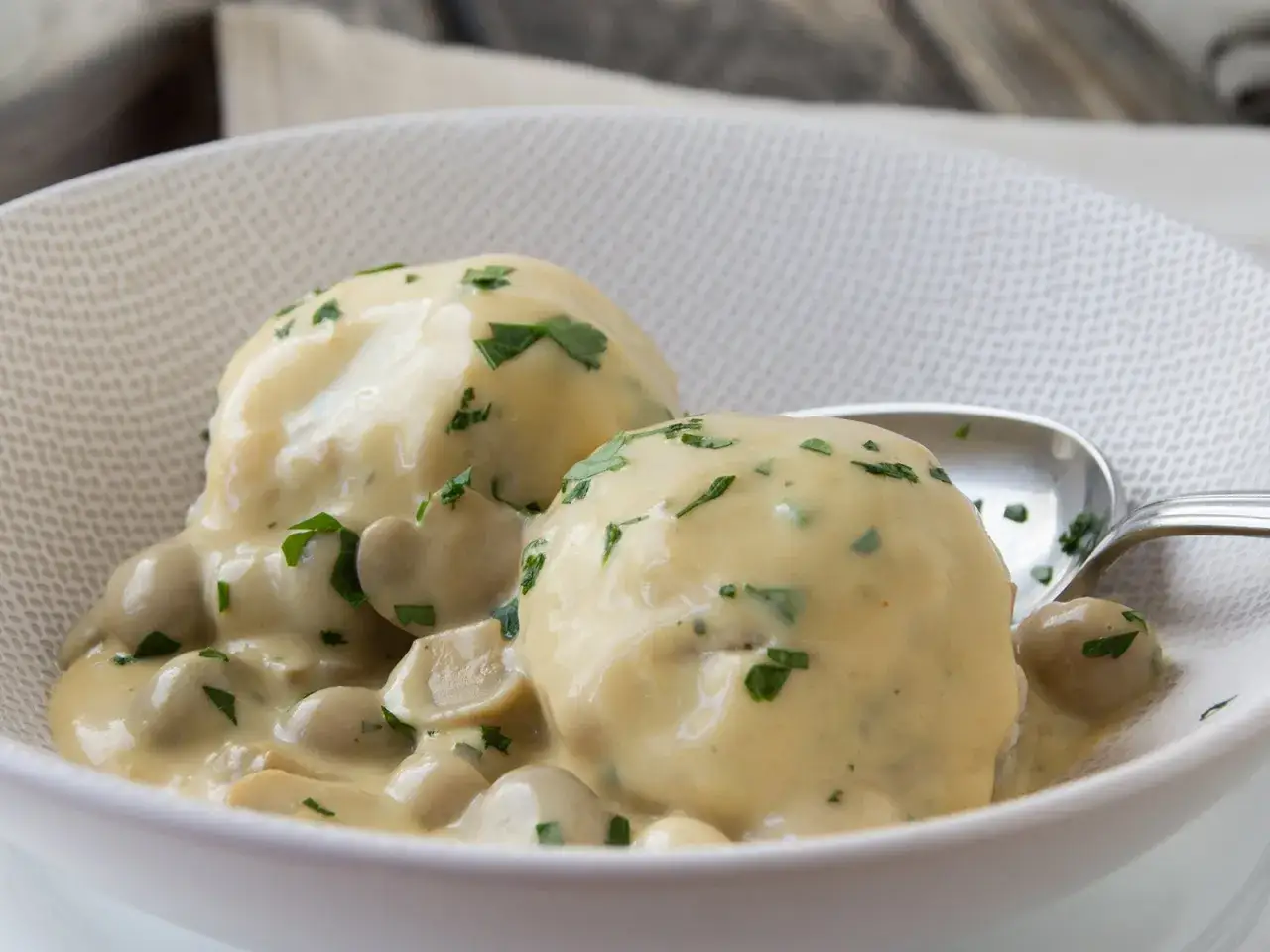 Zwei Klöße in cremiger funghi sauce mit Petersilie garniert, serviert in einer weißen Schüssel mit einem Löffel.