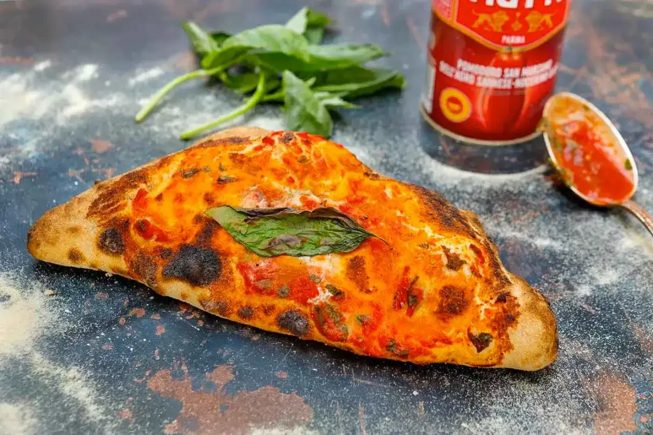 Ein knuspriger, original italienischer Calzone mit Tomaten und Basilikum, daneben eine Dose San Marzano Tomaten und ein Löffel Sauce.