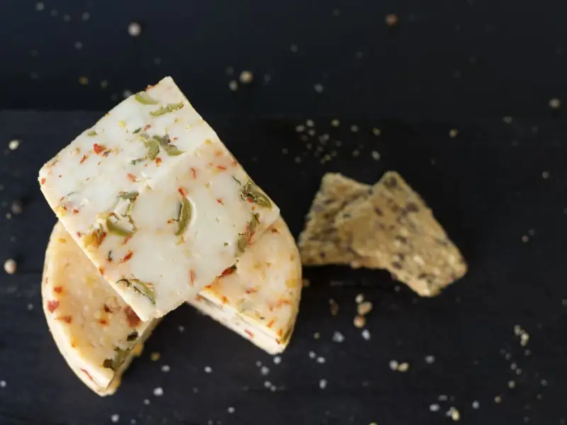 Käse mit Kräutern und Paprika, eine köstliche pecorino alternative, neben knusprigen Crackern auf dunklem Untergrund.