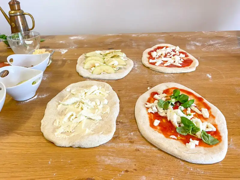 Vier vorbereitete Pinsa-Teiglinge liegen auf einem Holztisch. Einer mit Apfelscheiben, einer mit Tomatensoße und Mozzarella, einer nur mit Mozzarella und einer mit Tomatensoße, Mozzarella und Basilikum.